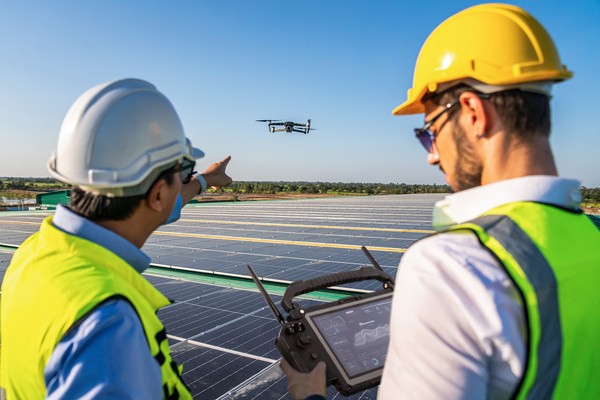 Two workers in hard hats and safety vests operate a drone above rows of solar panels on a solar farm.